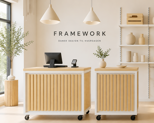 Butiksdisk i lyst træ med hvid ramme og hjul, med kasseskærm og betalingsterminal, foran en væg med teksten "Framework"